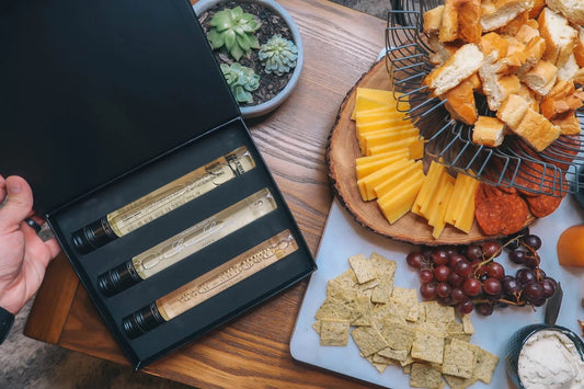 Gift box with three wine bottles beside a charcuterie board of cheese, meats, bread, grapes, and crackers.