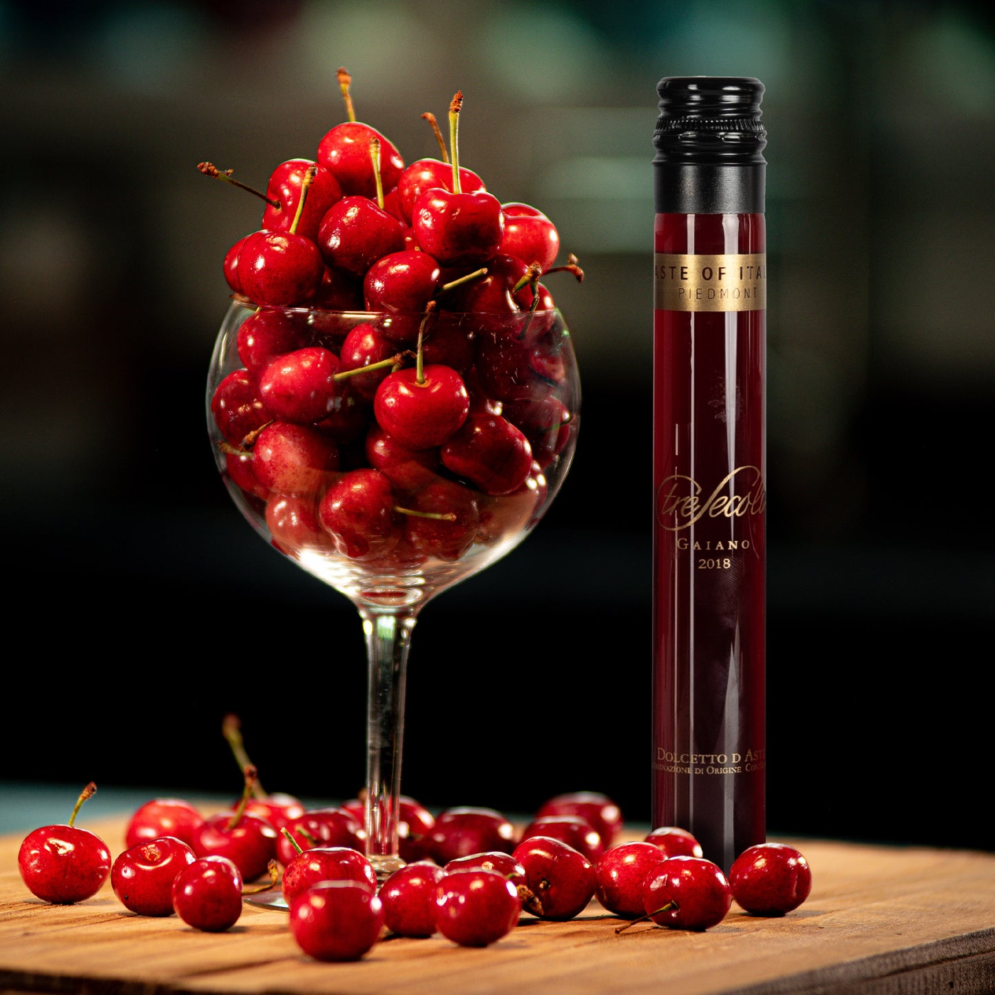 Glass filled with cherries next to a bottle of red wine on a wooden surface.