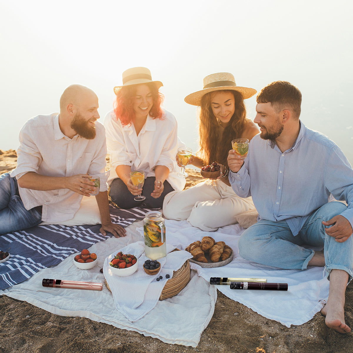 Four friends enjoying a picnic on a sandy beach with drinks and snacks.