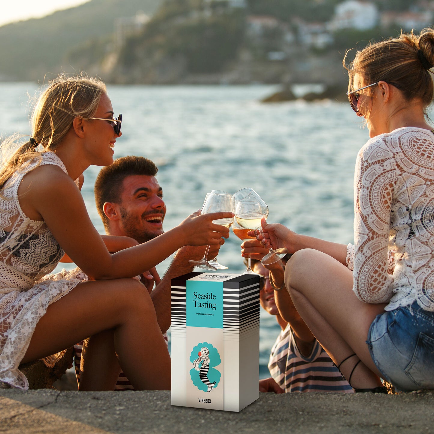 Two women and a man sitting by a body of water, enjoying wine and a box labeled 'Seaside Tasting'.