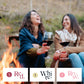Two women sitting by a campfire holding wine glasses